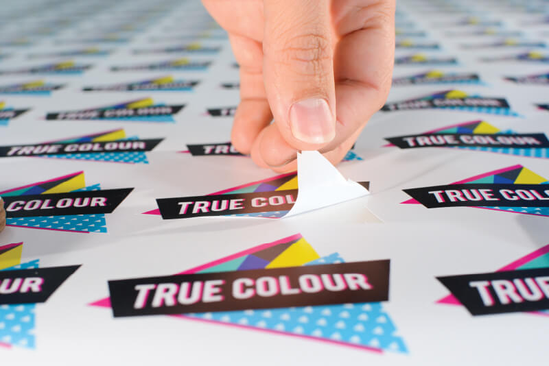 A hand peeling a full-colour sticker off a sheet of many stickers