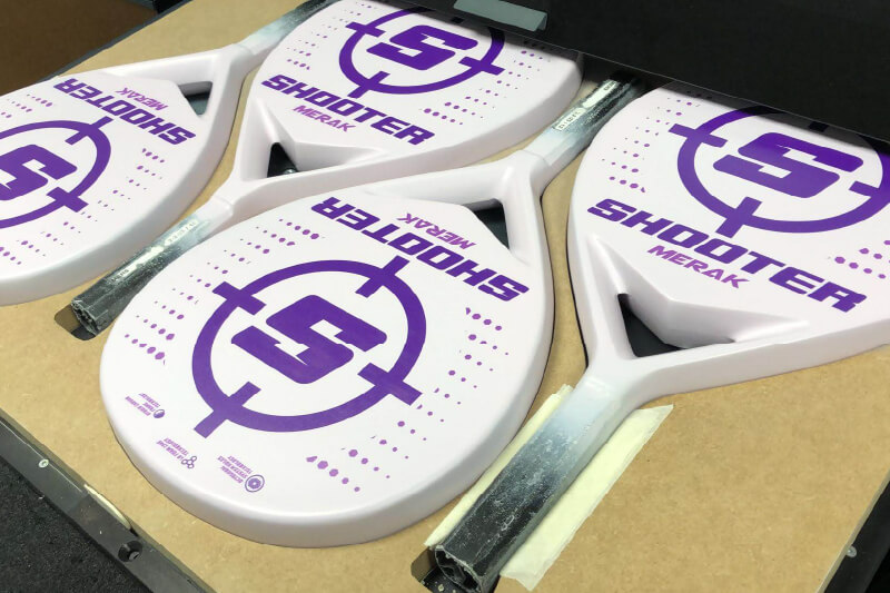 Four custom printed white padel rackets secured in a jig on a flatbed UV printer.